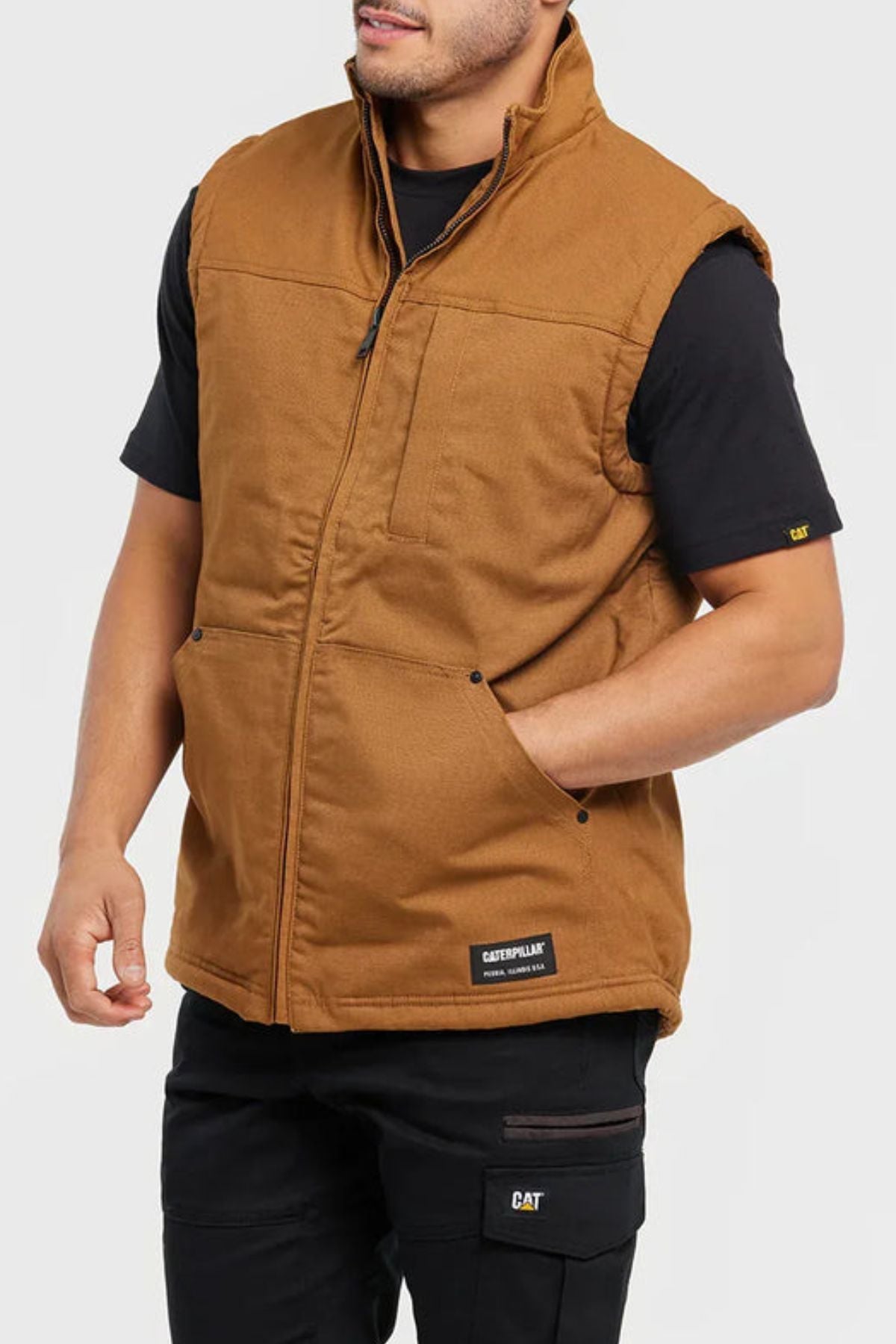 A man wears the CAT Sherpa Lined Vest—a brown, sleeveless, insulated vest with front zipper, chest and hand pockets, and water-resistant finish—over a black CAT T-shirt and pants. The background is plain light gray.
