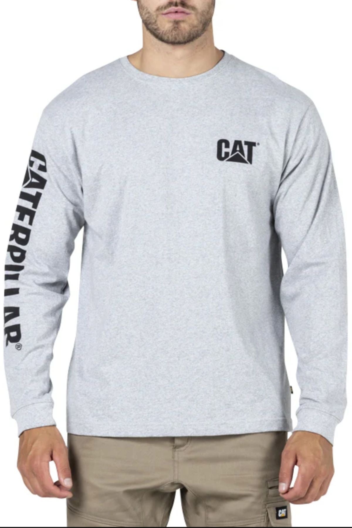 A man wears a CAT Trademark Banner Long Sleeve Tee featuring the CAT logo on the chest and "CATERPILLAR" down the left sleeve, paired with khaki pants, and stands facing forward.