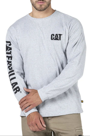 A man wears the CAT Trademark Banner Long Sleeve Tee in light gray, featuring a "CAT" logo on the chest and "CATERPILLAR" on the right sleeve, paired with tan pants for a comfortable, relaxed everyday look.