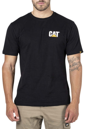 A man stands facing forward, wearing the CAT Trademark Tee in black, featuring a small left chest logo. The 100% cotton crew neck t-shirt is paired with beige pants. Only his upper body is visible; he has a right forearm tattoo.