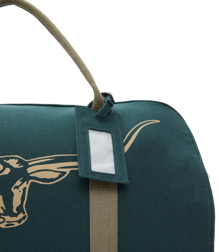 A close-up of the RM Williams Nanga Canvas Bag in dark green with a tan handle, rectangular luggage tag, and a partial long-horn steer head illustration on its durable cotton canvas exterior.