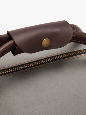 Close-up of the RM Williams Canvas Ute Bag highlighting a dark brown leather handle with a "Made in USA" brass button. The water-resistant light gray utility canvas features a visible brown zipper and tan stitching.