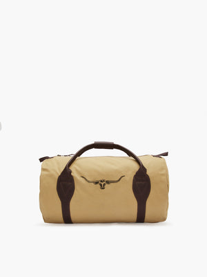 The RM Williams Canvas Ute Bag is a beige cylindrical bag made from water-resistant canvas, featuring dark brown leather handles and accents. It sports an embroidered longhorn skull logo on the front, combining simplicity and ruggedness—ideal for travel or gym use.