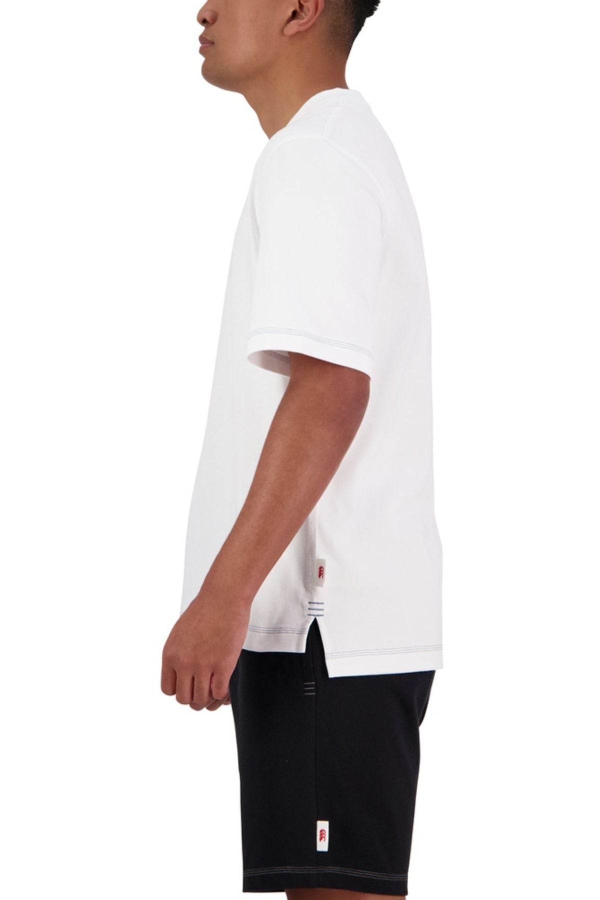 A person stands sideways in an oversized Canterbury CCC Collection T-Shirt in plain white and black shorts, set against a plain white background.