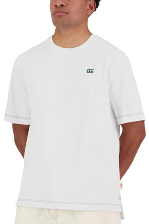 A person wears the Canterbury CCC Collection T-Shirt, a plain white tee with a small green logo on the left chest. Designed for comfort, it features an oversized fit and is shown with hands behind their back against a white background.