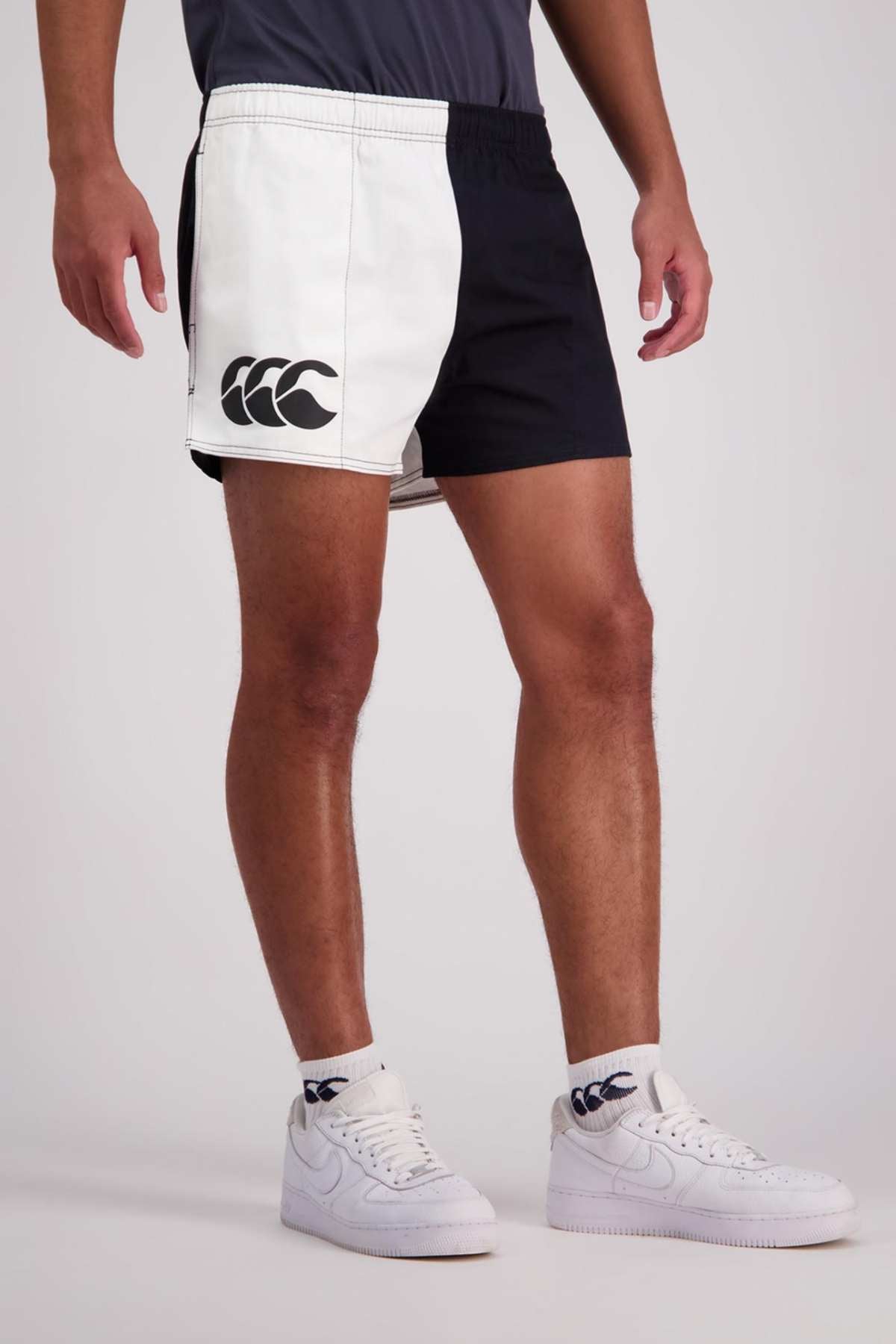 A person stands wearing the Canterbury Cotton Twill Harlequin Short, showcasing its distinctive black and white design with a large logo on the left leg. They also sport white sneakers and white socks, each adorned with a similar logo. These shorts provide a customizable fit and breathable comfort. The backdrop is plain gray.