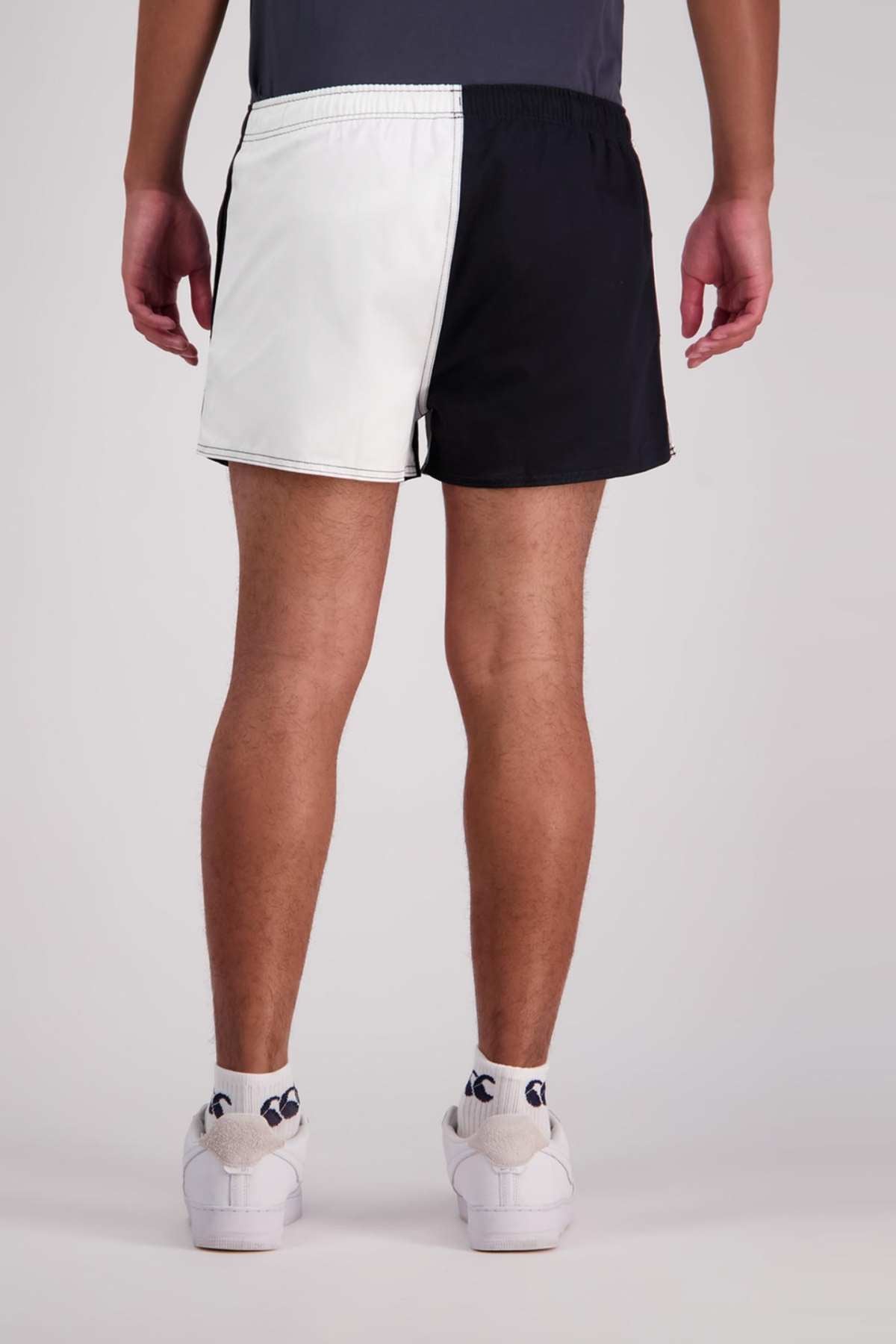 A person, seen from behind, is wearing Canterbury Cotton Twill Harlequin Shorts, white socks with a small black design, and white sneakers. They also have on a dark gray shirt with its hem just barely visible. The background is solid gray.