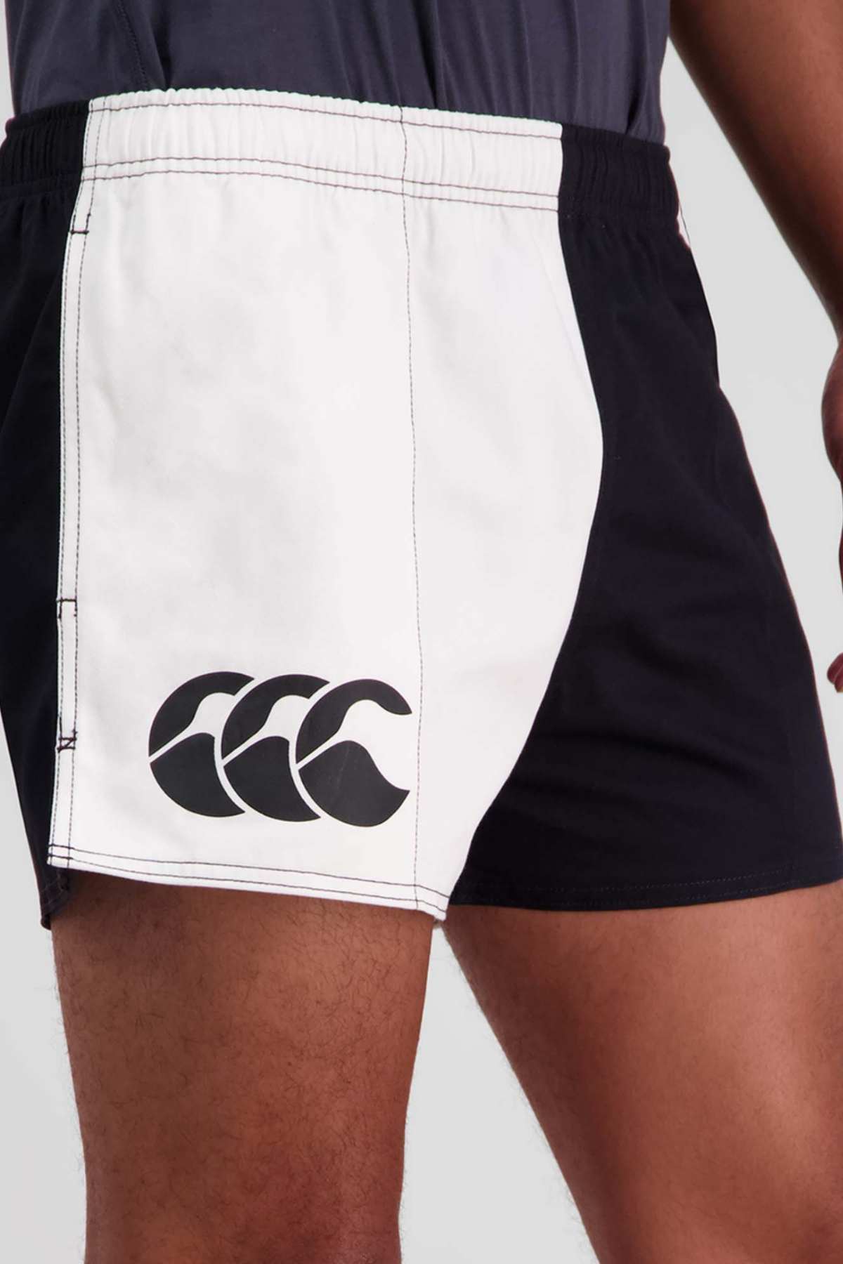 A person wearing Canterbury Cotton Twill Harlequin Shorts in black and white, featuring a graphic logo on the left side. The person's hand is resting on their hip as they showcase the breathable comfort of the shorts, set against a plain, neutral background.