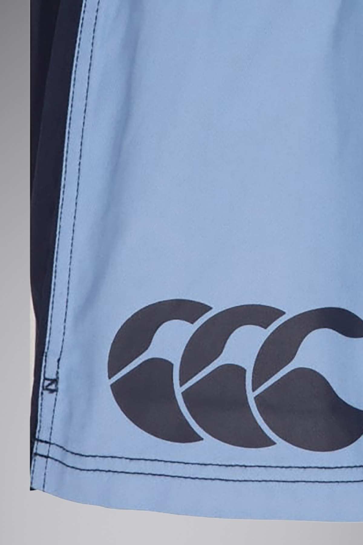 Close-up view of a garment made of light blue 100% cotton twill fabric with black stitching along the edge. The fabric features a black logo with three stylized overlapping waves near the bottom, reminiscent of the Canterbury Cotton Twill Harlequin Short design.