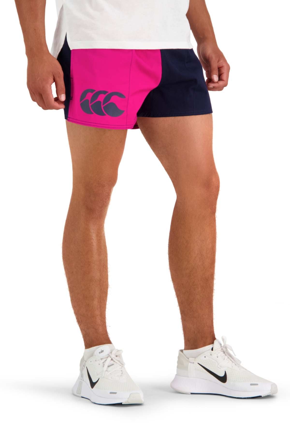 A person is pictured from the waist down wearing a white t-shirt, Canterbury Cotton Twill Harlequin Short with pink and black panels offering breathable comfort, and white Nike sneakers. The logo on the shorts features three overlapping outlines. The person is standing on a white background.