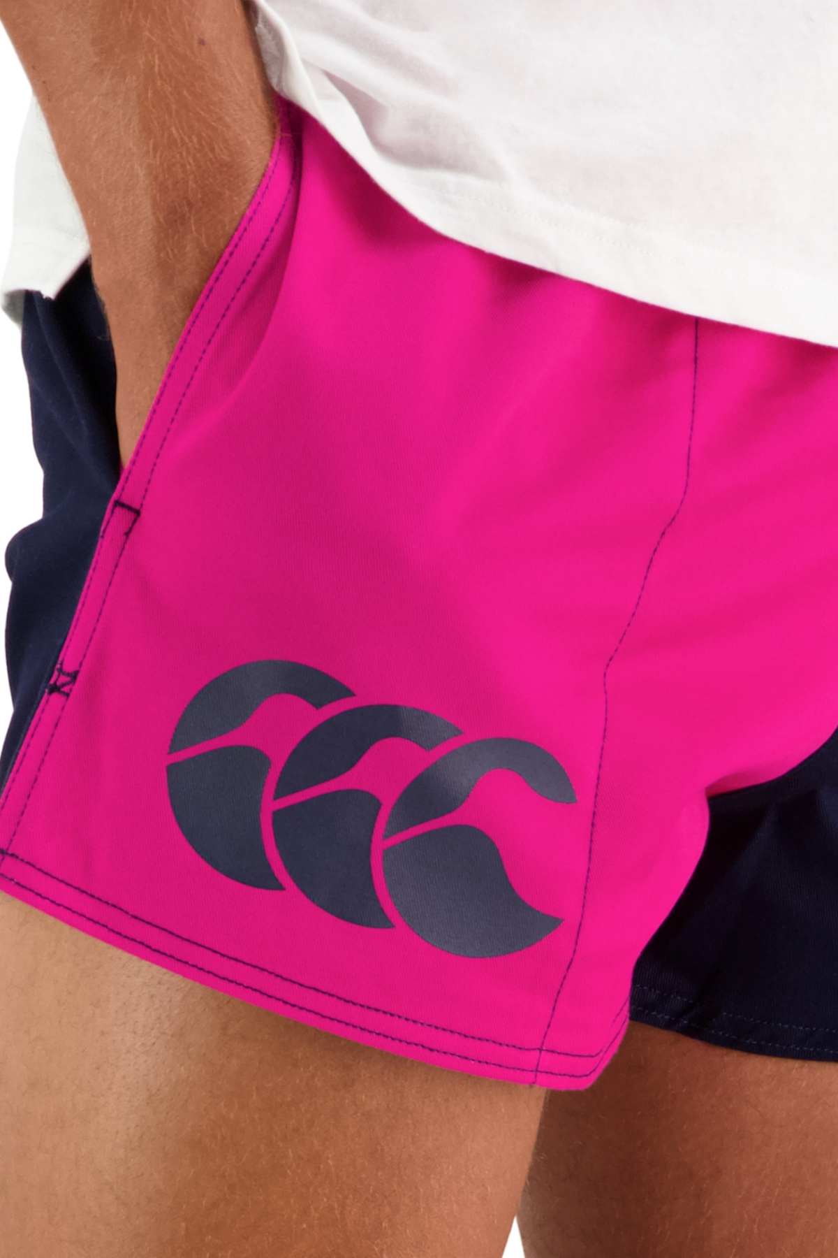 A person is wearing a pair of bright pink Canterbury Cotton Twill Harlequin Shorts, featuring a navy blue logo on the side pocket. These shorts provide a customizable fit and breathable comfort, making them ideal for both athletic and casual wear. The individual is partially visible with one hand resting in the pocket of the shorts.