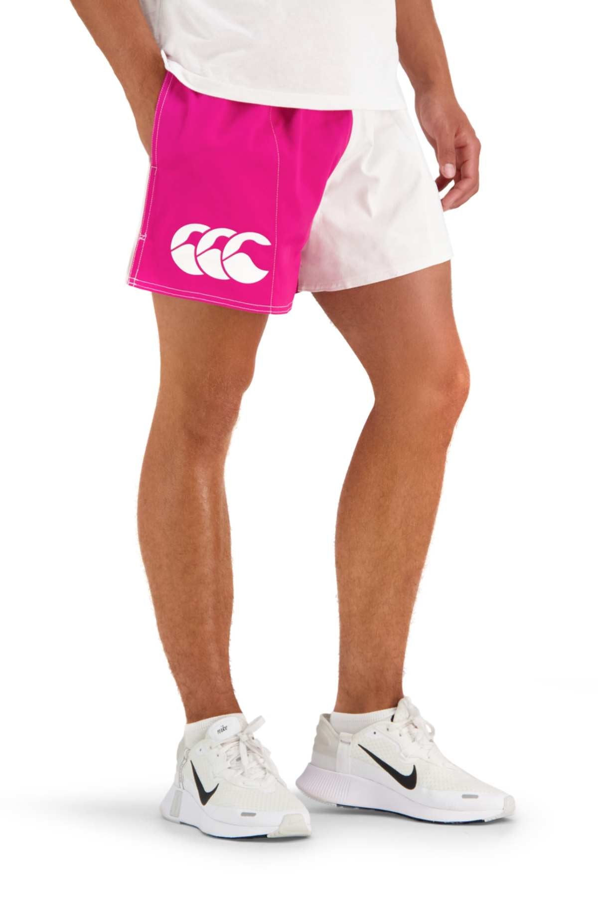 A person wears a white T-shirt, Canterbury Cotton Twill Harlequin Shorts in bright pink and white with a large white logo, and white Nike sneakers with black swooshes, standing against a white background.