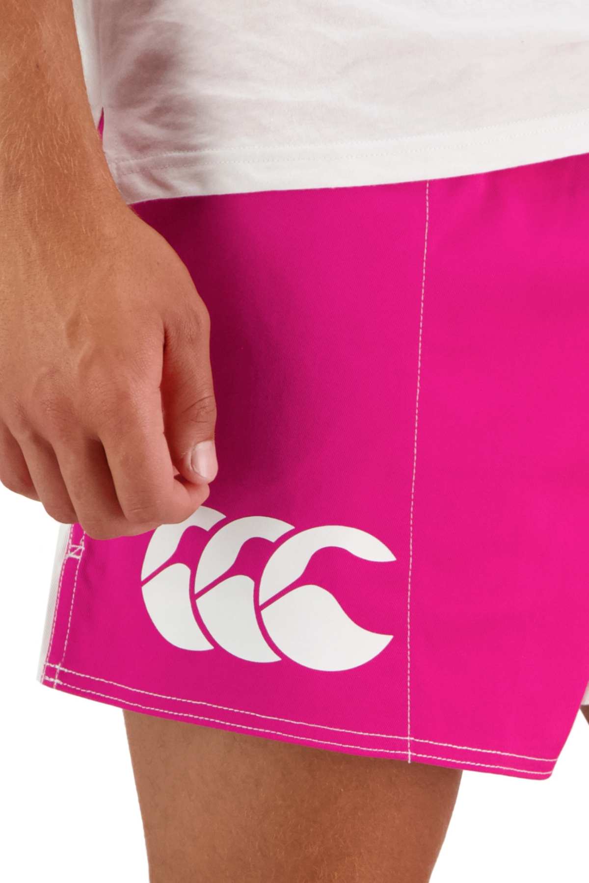 A person wears bright pink Canterbury Cotton Twill Harlequin Shorts with a white logo, standing with their arm relaxed at their side. The background is plain, and the upper part of their white shirt is partially visible.