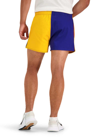 A person wearing a white T-shirt and the brightly colored Canterbury Cotton Twill Harlequin Short, featuring one leg in yellow and the other in blue, paired with white sneakers. The individual is shown from behind, highlighting the back view of the outfit that assures breathable comfort.
