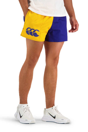 A person is dressed in Canterbury Cotton Twill Harlequin Shorts in yellow and blue, paired with a white shirt. The breathable shorts display the Canterbury logo, featuring three stylized, overlapping shapes. Completing the outfit are white athletic sneakers with a black swoosh logo. The background is plain white.