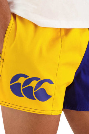 Close-up view of a person wearing yellow and blue Canterbury Cotton Twill Harlequin Shorts with an elasticated waist and a blue wave-like logo on the left side. The person's white shirt is partially visible at the top of the image, emphasizing breathable comfort.