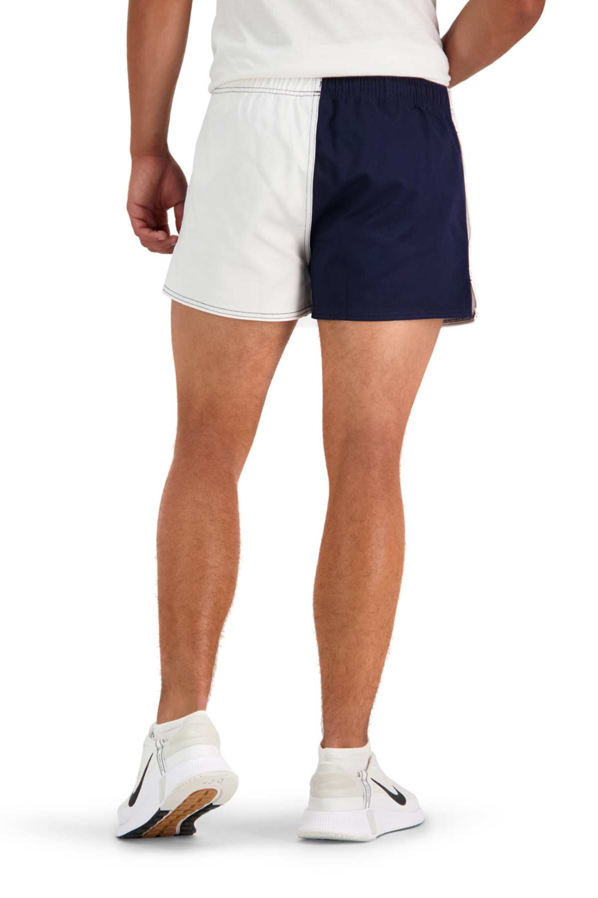 A person in a white t-shirt is wearing Canterbury Cotton Twill Harlequin Shorts, featuring one side in white and the other in dark blue. They are also sporting white athletic shoes with black swoosh logos and fluorescent green accents, designed for breathable comfort. The person is standing with their back to the camera.