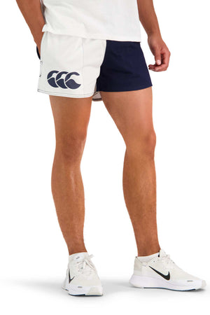 A person wearing a white t-shirt, Canterbury Cotton Twill Harlequin Short featuring a large logo on the left leg, and white Nike sneakers is standing against a plain white background. The breathable comfort of the shorts complements the relaxed posture as their arms rest casually by their sides.