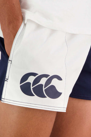 Close-up of a person wearing the Canterbury Cotton Twill Harlequin Short, white athletic shorts with blue accents. Known for their breathable comfort and customizable fit, these shorts feature a navy blue logo with three stylized waves on the left side. The person's hand is resting near the pocket of the shorts.