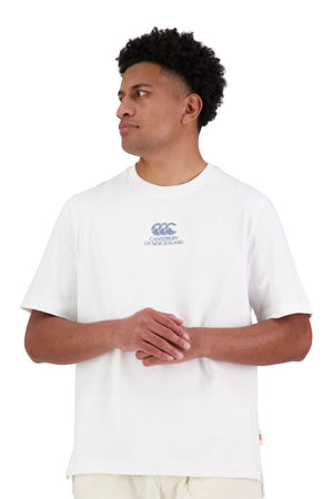 A man wearing the Canterbury Heritage T-Shirtβan oversized, heavy-duty 100% cotton white tee with a small blue CCC logo on the chestβstands against a white background, looking to his left with his hands loosely clasped.