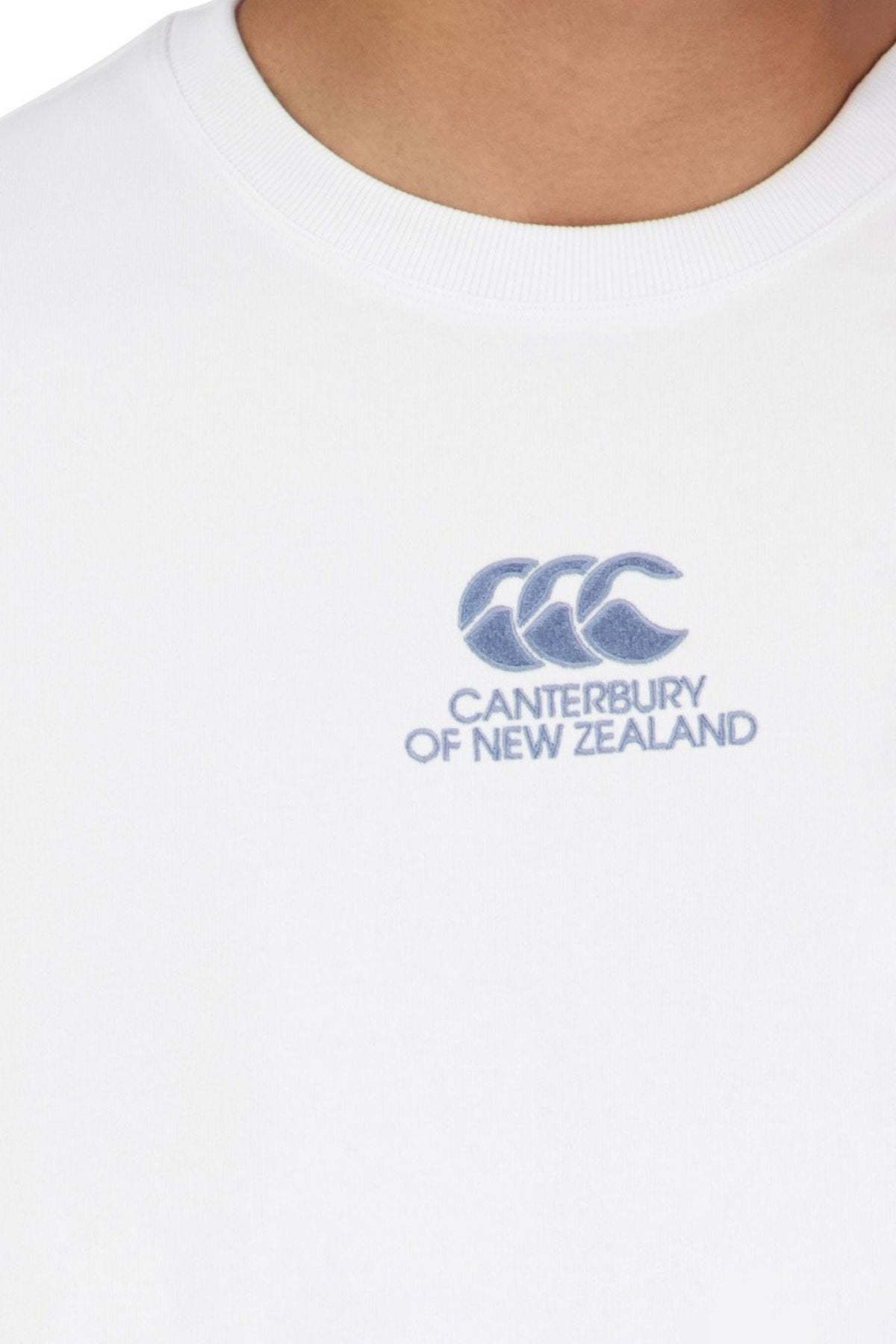 Close-up of a person wearing the Canterbury Heritage T-Shirt, featuring the CCC logo and "Canterbury of New Zealand" embroidered in blue on the chest. Only the lower neck and upper chest are visible.