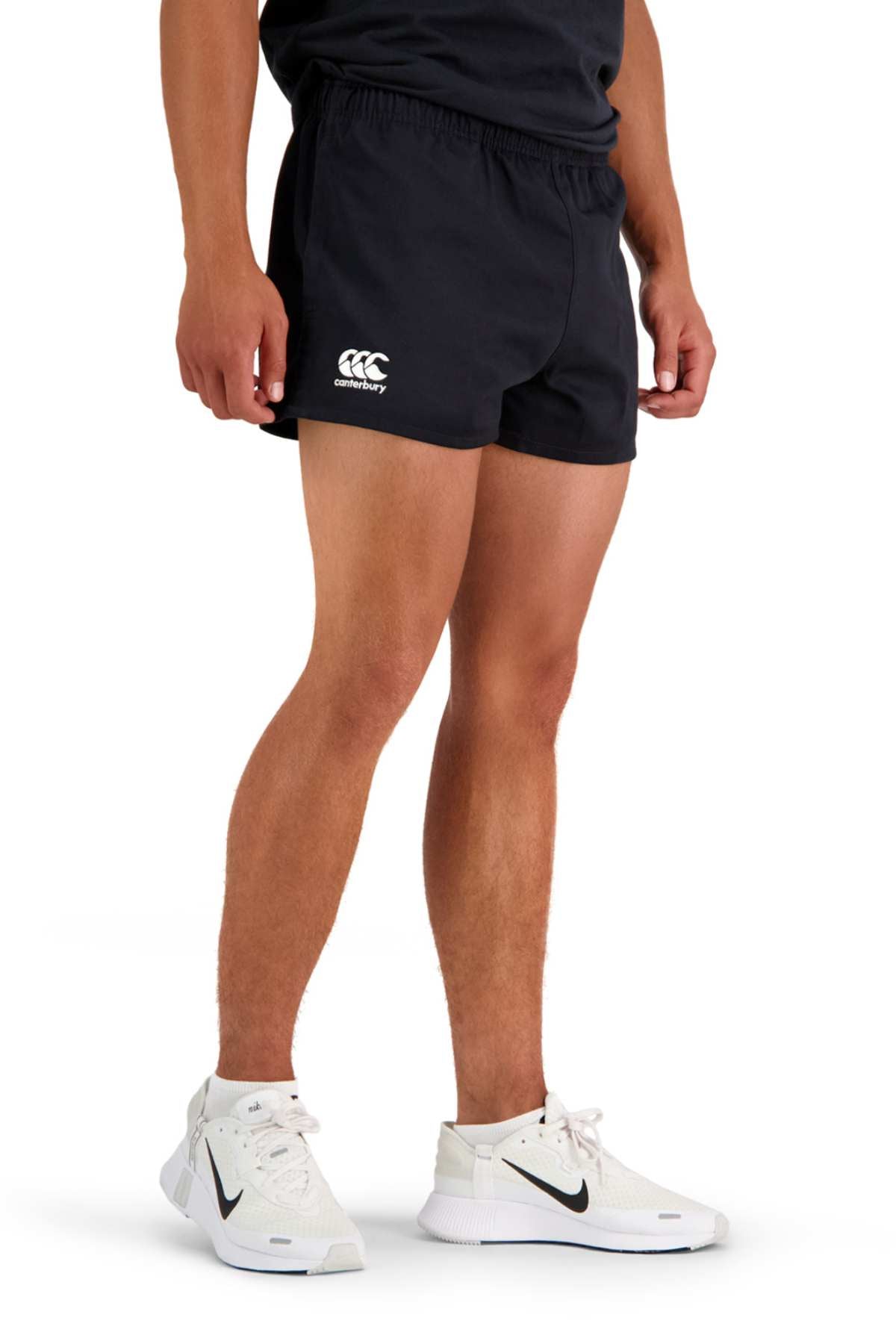 A person is standing in a relaxed pose, shown from the torso down, wearing black Canterbury Rugged Drill Shorts with an elasticated waist and a white Canterbury logo on the left leg. They are also wearing a black top, white high-top sneakers featuring a black Nike logo, and white socks.
