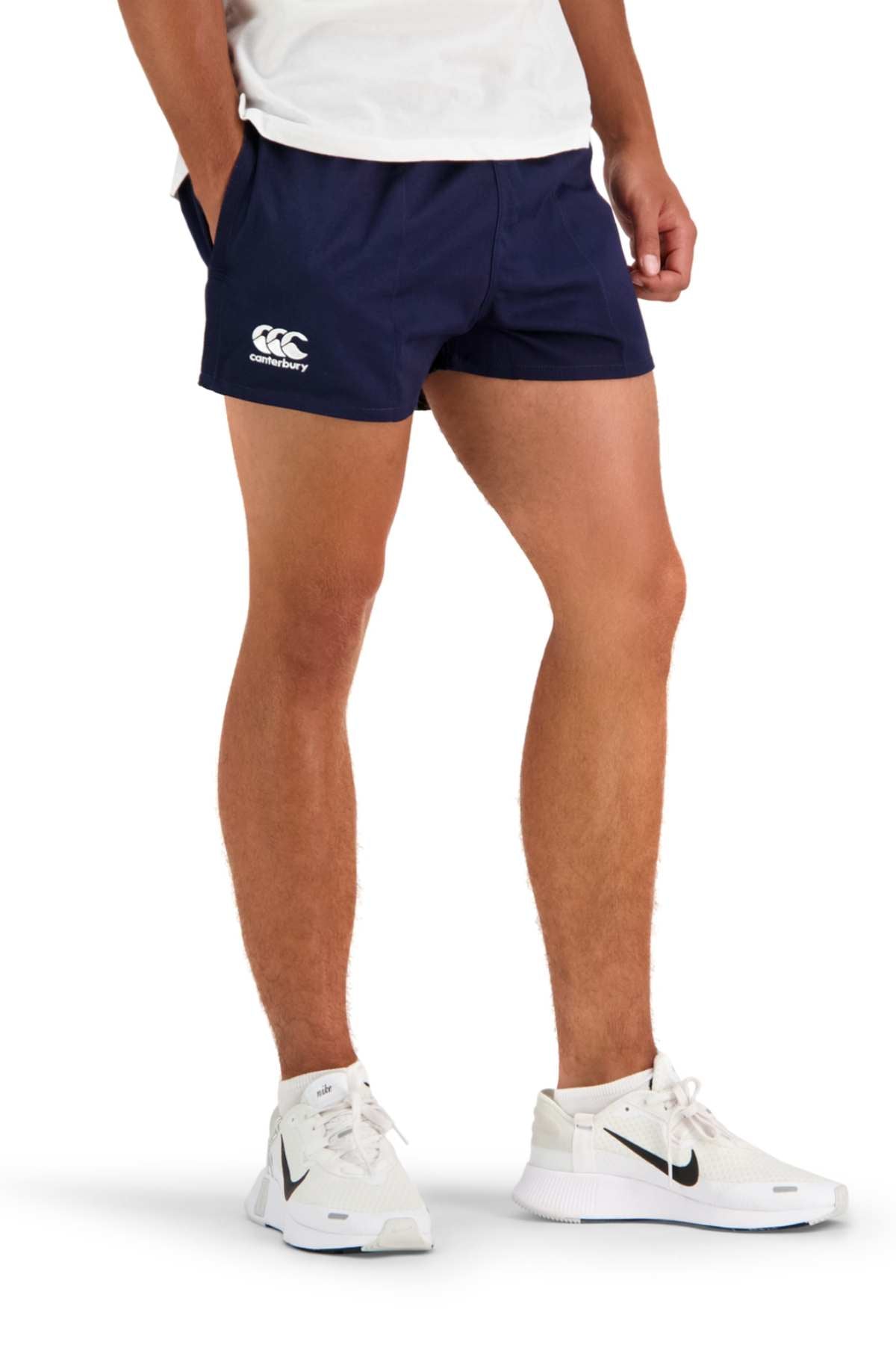 A person is wearing a white shirt, navy blue Canterbury Rugged Drill Shorts with an elasticated waist, and white Nike sneakers. The individual stands casually with hands in pockets, showcasing a sporty look focused on the lower half of the body.