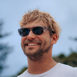 A smiling man with tousled blond hair and light facial hair stands outdoors in a white t-shirt, wearing SafeStyle Classics Graphite Frame Polarised Lens sunglasses. The background of trees and blue sky is blurred.