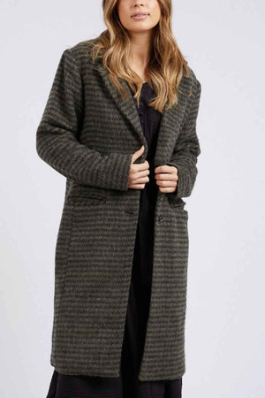 A woman with long hair is wearing the Foxwood Clementine Coat, a green and black striped, midi-length jacket made from a wool blend. The coat has buttons and two side pockets. Standing against a plain background, she lightly grips the edges of her coat, revealing her black top underneath.