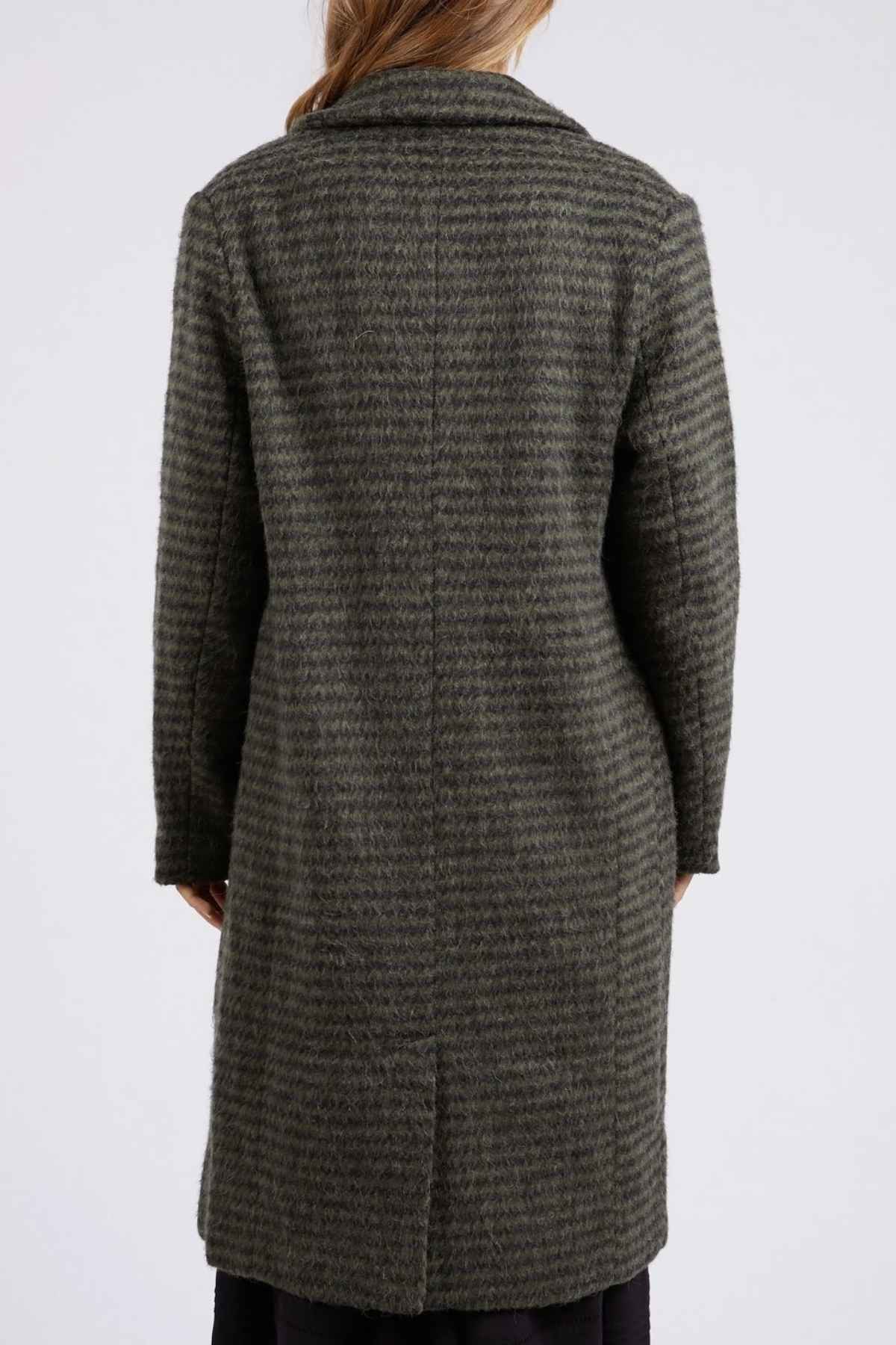 A person with long hair, seen from the back, is wearing a Foxwood Clementine Coat featuring dark green stripes that extends to just above the knees. The coat, crafted from a wool blend with a textured pattern and a single vent at the back, stands out against the plain white background.