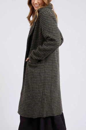 A person is dressed in a long, textured dark green Foxwood Clementine Coat that features a subtle striped pattern. They are standing sideways with one hand in their pocket and a slight smile on their face, looking at the camera against a plain and light-colored background.