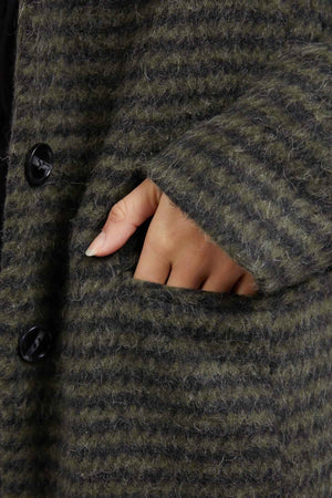 A hand is partially inserted into the pocket of the Foxwood Clementine Coat, a finely textured wool blend coat in dark green and black stripes. The close-up shot highlights its visible black buttons while focusing on the pocket area and fingers.