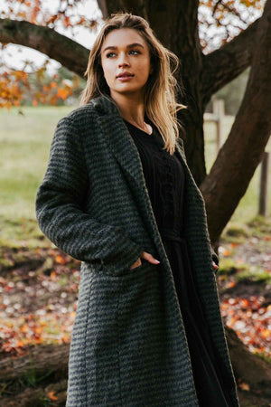 A person with long hair stands outdoors, looking thoughtfully into the distance. They are wearing the Foxwood Clementine Coat featuring a cozy wool blend and a stylish check pattern, with their hands tucked into the coat’s pockets. Autumn leaves and a large tree are visible in the background.