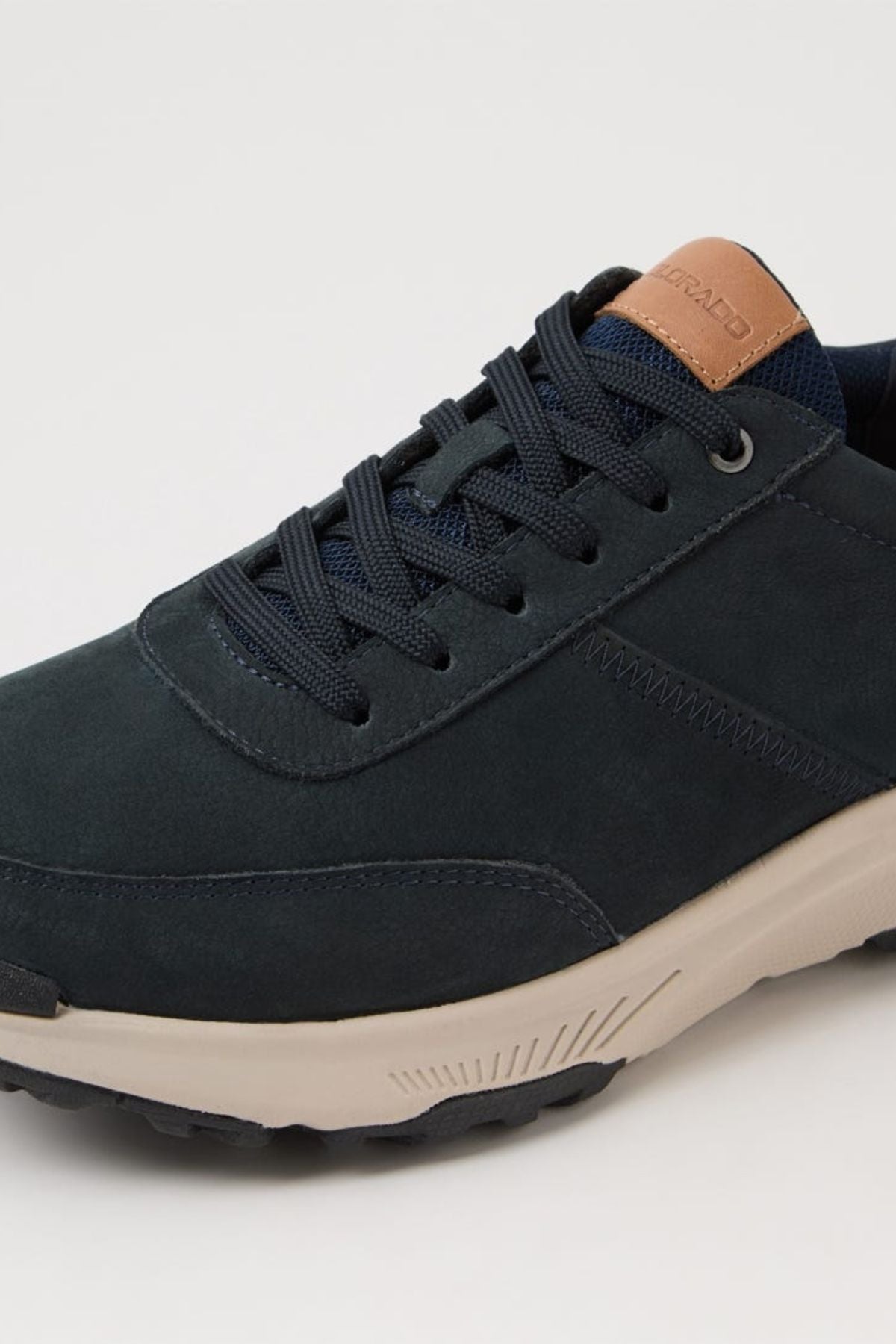 A close-up of the Colorado Suzuki Shoe reveals dark blue suede, navy laces, a beige slip-resistant sole, and a brown leather tongue accent with embossed text.