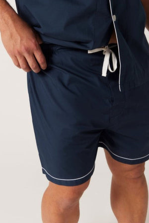 Donning the Contare Country Cotton Rich Short Sleeve Classic Set in navy blue with white trim, a person stands against a plain background. This stylish ensemble includes a short-sleeved top and drawstring shorts, emphasizing the contemporary look of their attire.