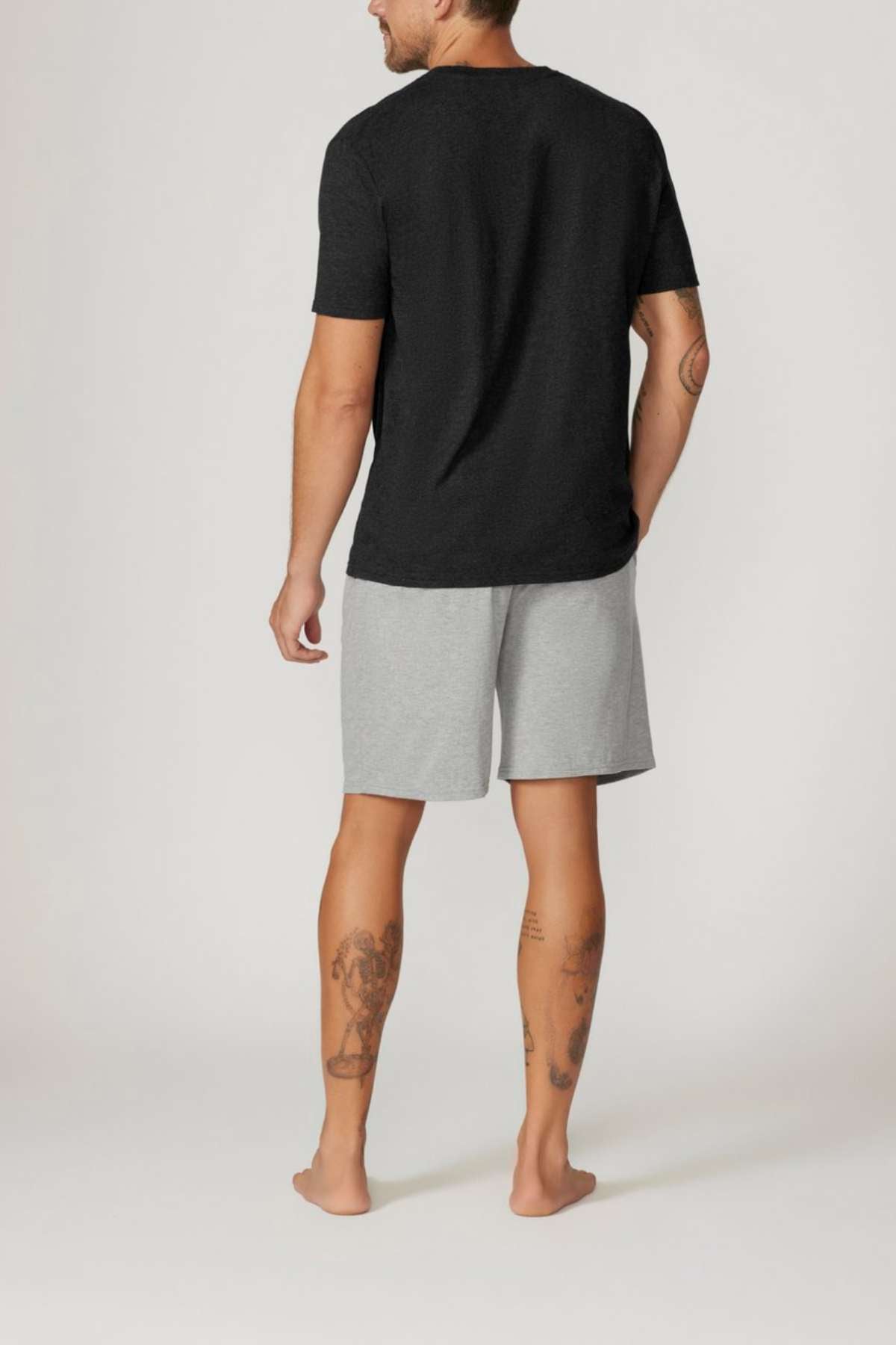 Facing away on a plain background, a person stands barefoot wearing the Contare Country Bamboo Cotton Short Sleeve Set. The set features a black T-shirt and gray shorts with hypoallergenic properties. Their tattooed arms and legs stand out against the simple backdrop.