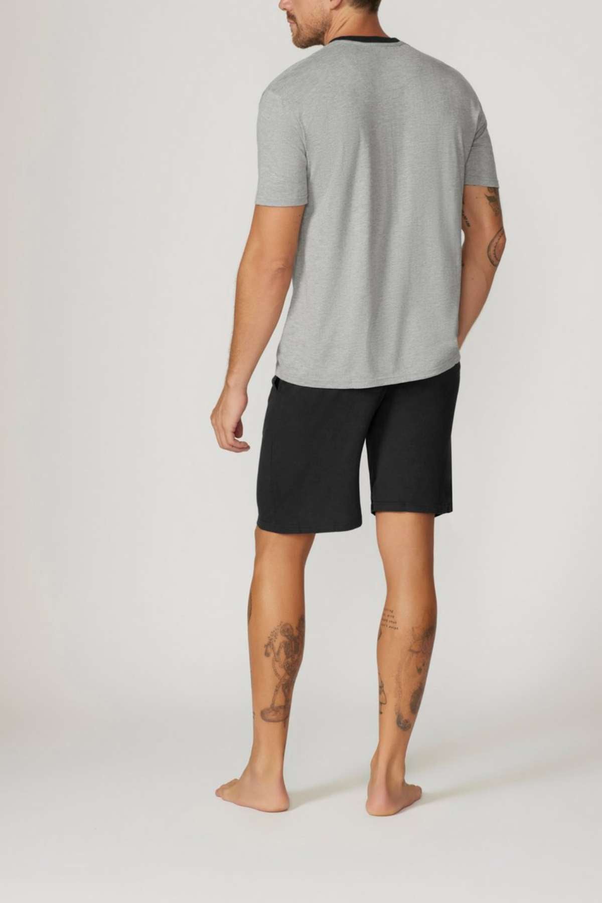 A barefoot tattooed man stands facing away, wearing the Contare Country Bamboo Cotton Short Sleeve Set in grey with black shorts, against a plain light background. The hypoallergenic set is crafted from bamboo cotton.