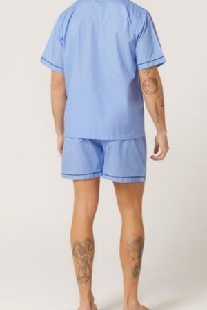 A person with tattoos on their arms and legs is standing barefoot, shown from the back, wearing the Contare Country Cotton Rich Short Sleeve Classic Set in light blue.
