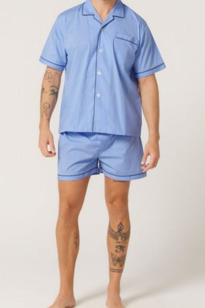 A person with tattoos on their arms and legs is wearing the Contare Country Cotton Rich Short Sleeve Classic Set—light blue pajamas with matching shorts—standing barefoot against a plain light background.