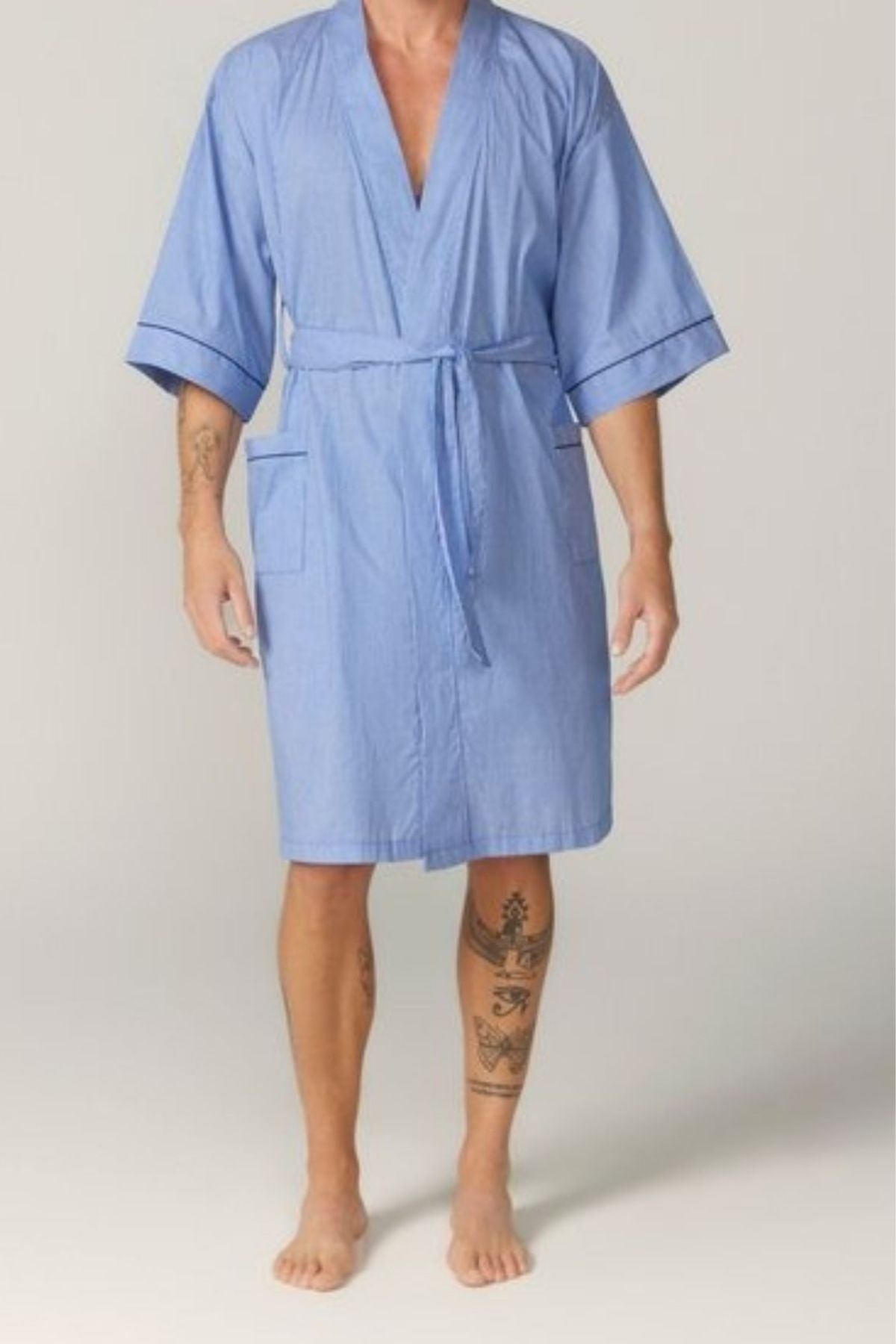 A person stands barefoot in the Contare Country Summer Dressing Gown, a light blue cotton kimono-style robe with short sleeves, front pockets, and a waist tie that falls above the knees, revealing tattoos on their left arm and right leg.