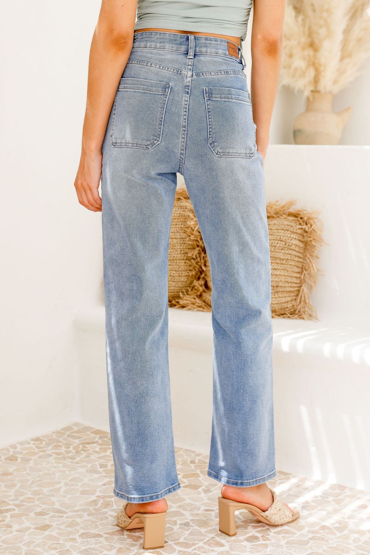 A person wearing Country Denim Straight Leg Stretchy Jean in light blue and block-heeled sandals stands indoors on a tiled floor with their back to the camera; a woven cushion and vase appear in the background.