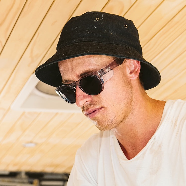 A person wearing a black bucket hat, SafeStyle Cruisers Graphite Frame Tinted Lens sunglasses with UV400 protection, and a white t-shirt is looking downward. The background features a light wooden ceiling. The expression appears focused or thoughtful.
