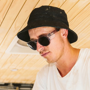 A person wearing a black bucket hat, SafeStyle Cruisers Graphite Frame Tinted Lens sunglasses with UV400 protection, and a white t-shirt is looking downward. The background features a light wooden ceiling. The expression appears focused or thoughtful.
