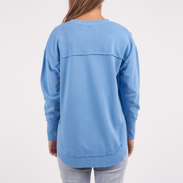 A person with long hair is seen from behind, wearing a light blue Foxwood Delilah Crew and matching jeans. The backdrop is a plain white canvas.