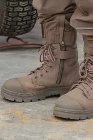 Close-up of a person wearing Steel Blue Argyle Zip Scuff Cap Boots with zippers and laces, paired with tan pants, standing on concrete. A large tire is visible in the background.