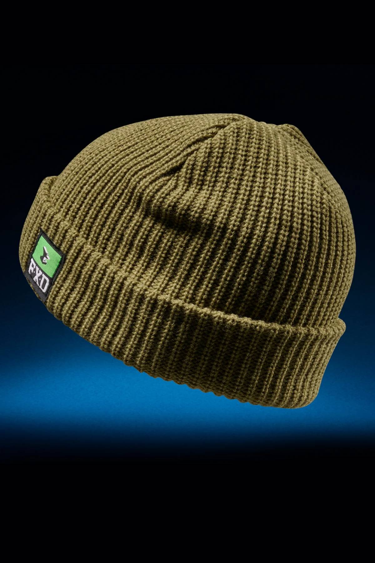 The FXD CP-10 Knit Ribbed Beanie is an olive green, double-layered 100% acrylic hat with a folded brim and a front patch featuring a green shield and "FXD" text, shown on a dark gradient blue background.