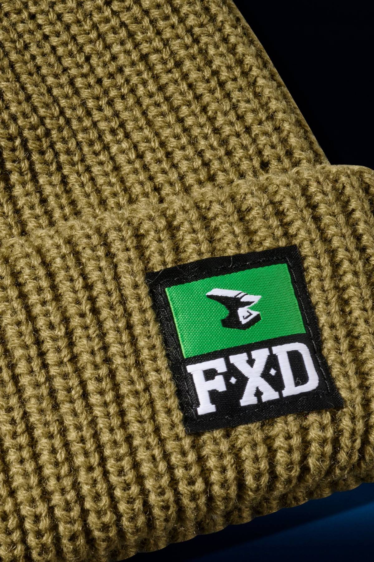 Close-up of the FXD CP-10 Knit Ribbed Beanie in olive green, made from 100% acrylic with a double-layer knit and folded brim, featuring a green and black patch with a white "FXD" logo on the front.