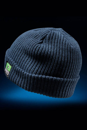 The FXD CP-10 Knit Ribbed Beanie is dark blue with a knit construction, a folded brim, and a small green and black rectangular patch on the front, shown against a dark gradient background.