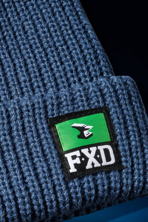 Close-up of the FXD CP-10 Knit Ribbed Beanie in blue with a black, green, and white FXD patch featuring a stylized bird logo on the turned-up brim and durable ribbed knit construction.