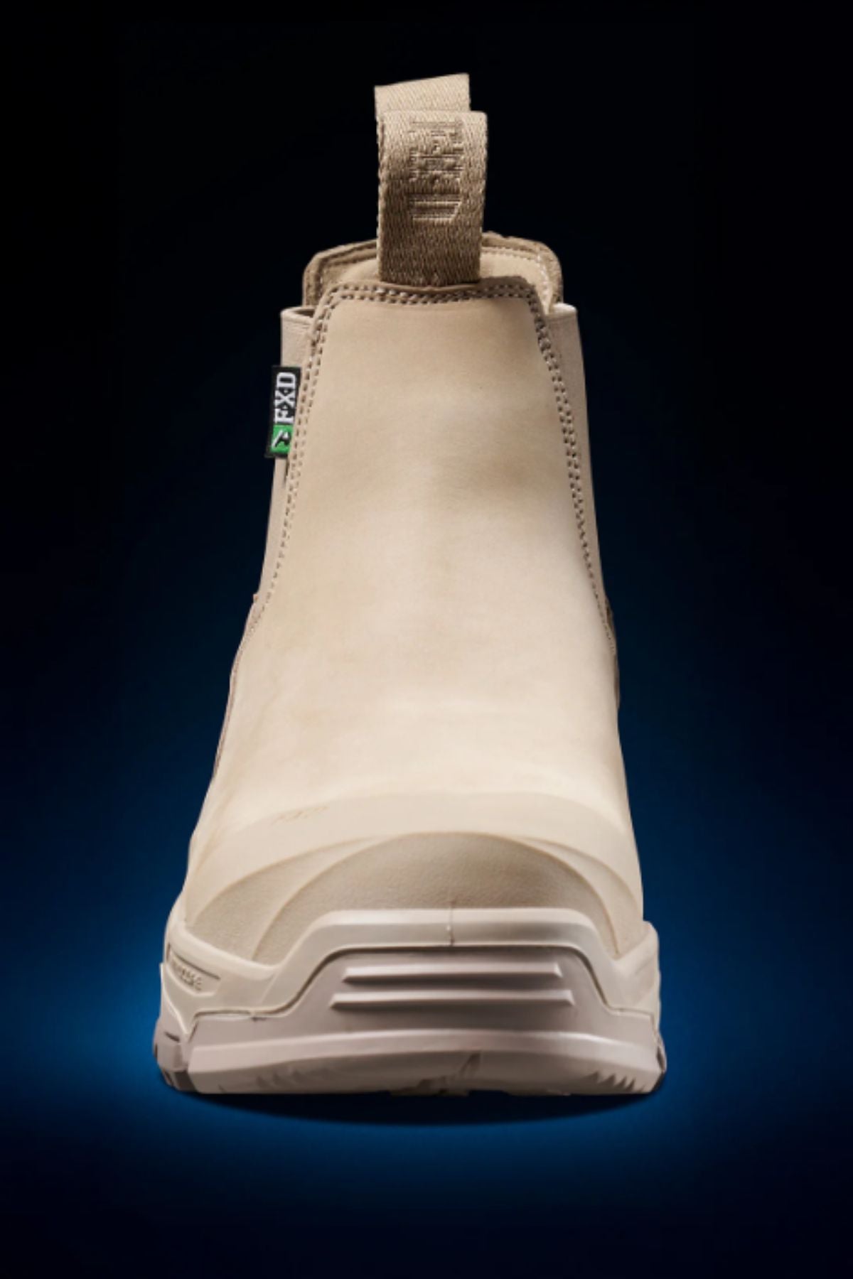 The FXD DURA X-3 Slip On Work Boots are shown from the front in tan, featuring a pull tab, reinforced toe, and rugged NITROCORE sole system, set against a dark gradient background.